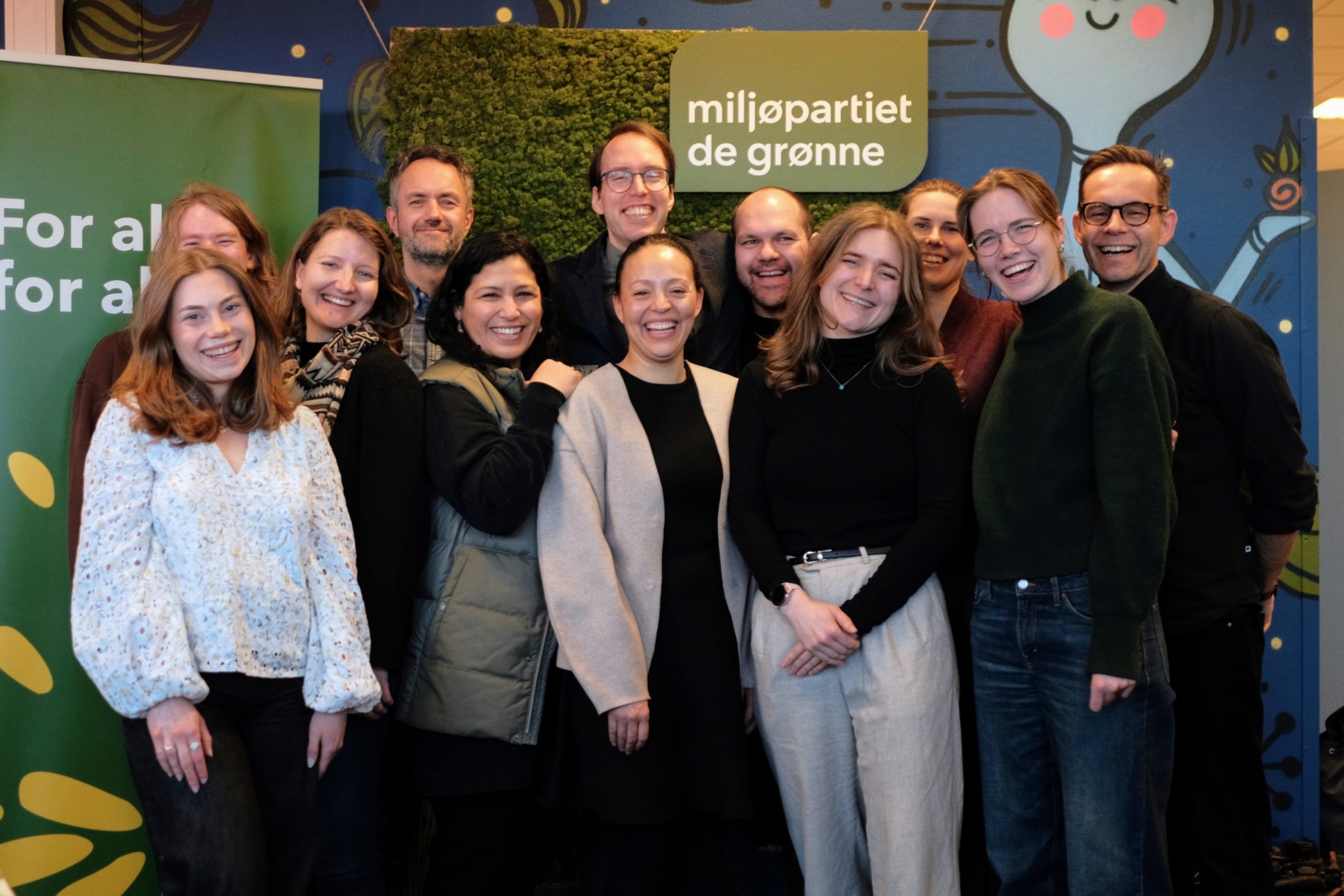 Styret i Oslo MDG, smilende foran plakater med logo og andre grønne budskap. Foto.